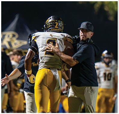 A person in a black shirt and cap is interacting with a football player wearing a yellow and white uniform with the number 2 on the back during a nighttime game. Descripci n generada con IA