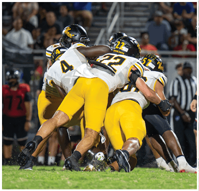 Several football players in yellow and white uniforms are tackling an opponent in a black and red uniform on a grassy field during a game. Descripci n generada con IA