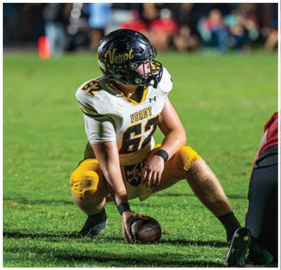 A person wearing a football uniform with a black helmet and yellow pants is crouched on a grassy field facing another player in a red and black uniform. Descripci n generada con IA