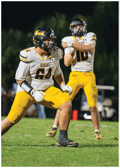 Two people in football uniforms with yellow pants and white jerseys are on a grassy field, with one person preparing to throw a football. Descripci n generada con IA