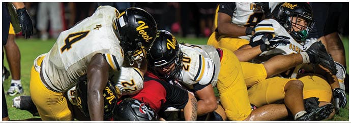 Several people wearing yellow and white football uniforms are engaged in a tackle during a game on a field. Descripci n generada con IA
