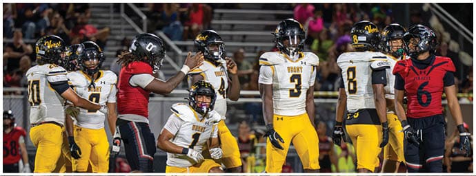 People wearing football uniforms in white and yellow or red and black are gathered on a field with stadium seating and spectators in the background. Descripci n generada con IA