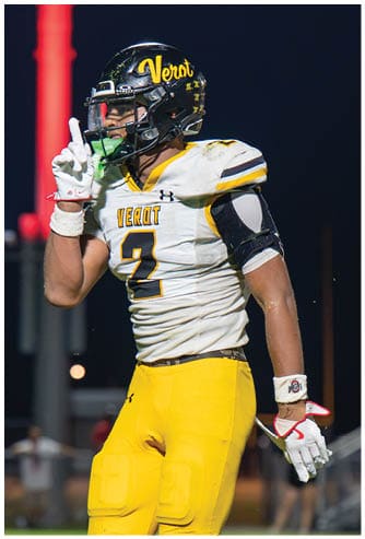 An adult wearing a football uniform with a helmet and gloves is raising one finger while standing on a field at night. Descripci n generada con IA
