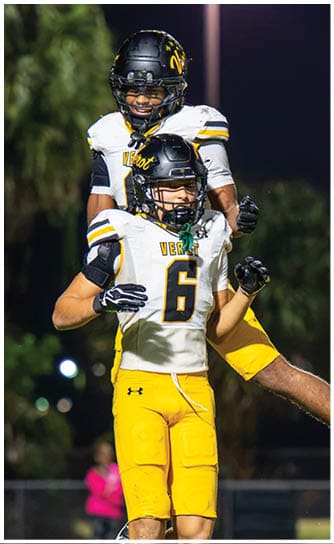 Two people wearing football uniforms with helmets are celebrating on a field at night, with one person lifting the other. Descripci n generada con IA