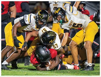 Several football players in yellow and white uniforms are tackling a player in a red and black uniform on a grassy field during a game. Descripci n generada con IA