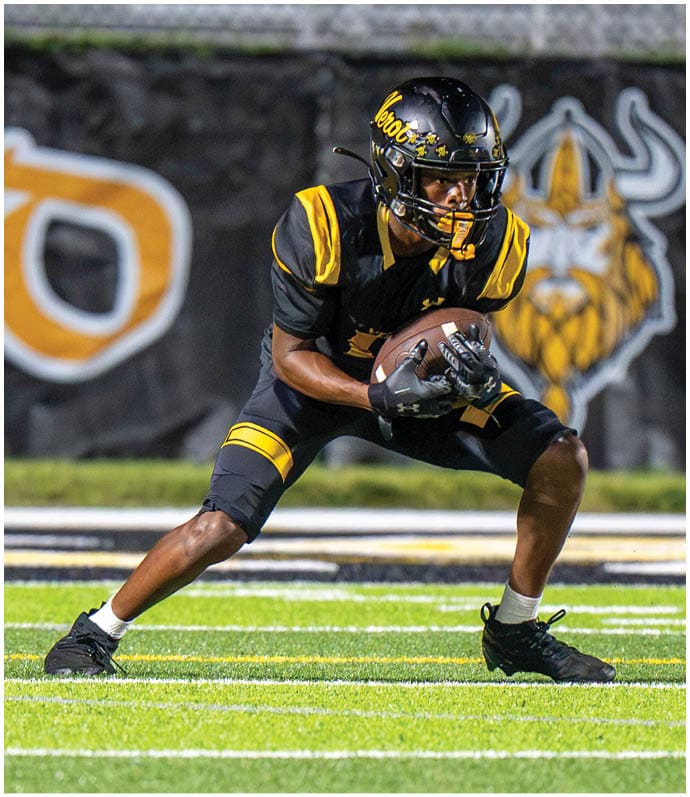 A person in black and yellow football gear is crouched on a green field holding a football, preparing for a play. Descripci n generada con IA