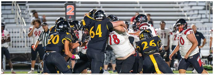 A group of people wearing football uniforms are engaged in a play on a football field with bleachers in the background. Descripci n generada con IA