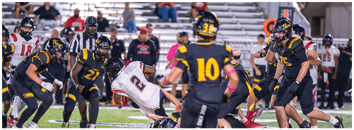 A group of people wearing football uniforms are actively engaged in a play on a football field with one person in white falling while others in dark uniforms surround them. Descripci n generada con IA
