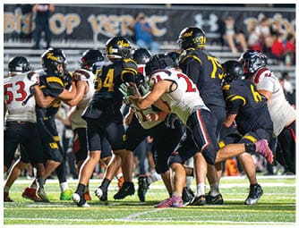 People wearing sports uniforms and cleats are engaged in a close physical play on a grassy field, likely during a football game. Descripci n generada con IA