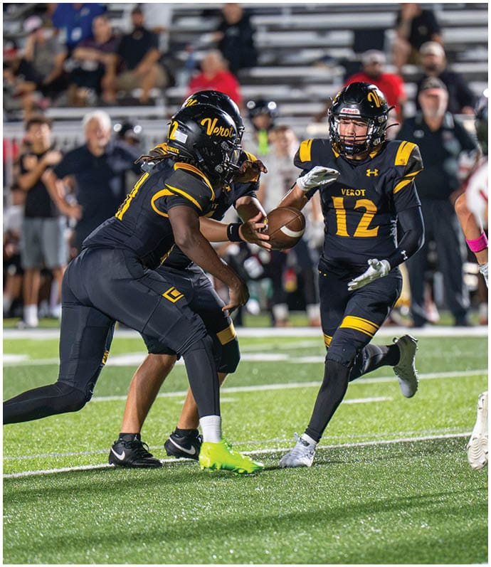 Two football players in black and yellow uniforms are engaged in a play on a field with one player handing the ball to the other while people watch from the stands in the background. Descripci n generada con IA