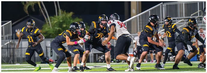 A group of people wearing football uniforms are engaged in a play on a well-lit field during a nighttime game. Descripci n generada con IA