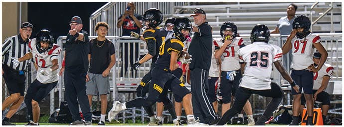 A group of people on a football field includes players in black and yellow uniforms and players in white and red uniforms, with some standing on the sidelines and others actively engaged in the game. Descripci n generada con IA