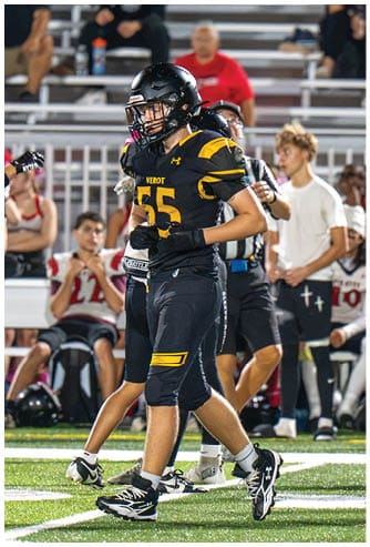 A person wearing a black and yellow football uniform with the number 55 is walking on a football field with other players and people in the background. Descripci n generada con IA