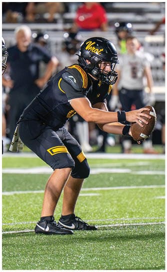 Three people in black and yellow football uniforms are on a green field with one person holding a football in a ready position while the others prepare to move. Descripci n generada con IA