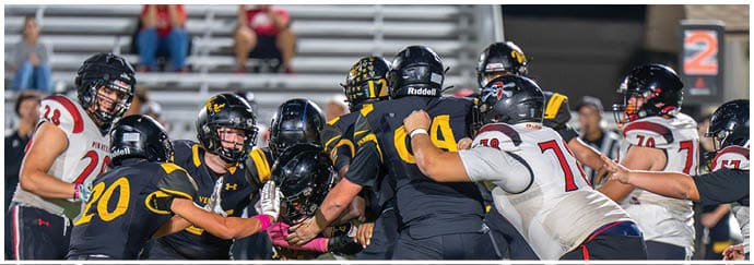 People wearing football helmets and uniforms are engaged in a close physical play on a football field during a game. Descripci n generada con IA