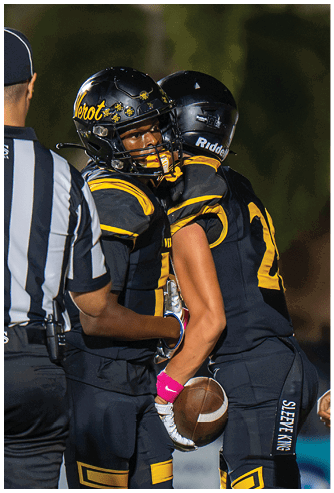 Two people in black and yellow football uniforms and helmets are embracing on a field with a blurred background. Descripci n generada con IA