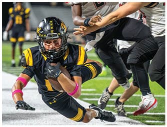 A person in a black and yellow football uniform is diving forward while holding a football tightly as two people in white and black uniforms attempt to tackle them on a football field. Descripci n generada con IA