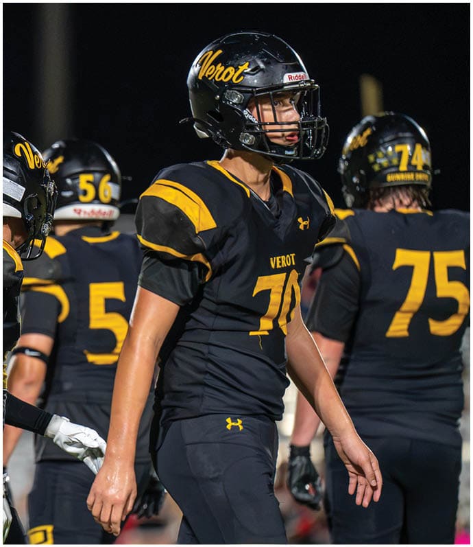 A person wearing a black and yellow football uniform and helmet with the word Verot stands on a field at night with other similarly dressed players in the background. Descripci n generada con IA