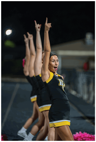 Adults in matching cheerleading uniforms perform a synchronized routine with one arm raised and fingers pointing upward against a dark background. Descripci n generada con IA