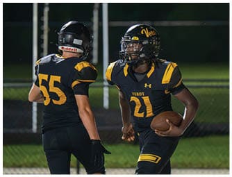 Two people in black and yellow football uniforms are on a field, with one holding a football and the other facing away. Descripci n generada con IA
