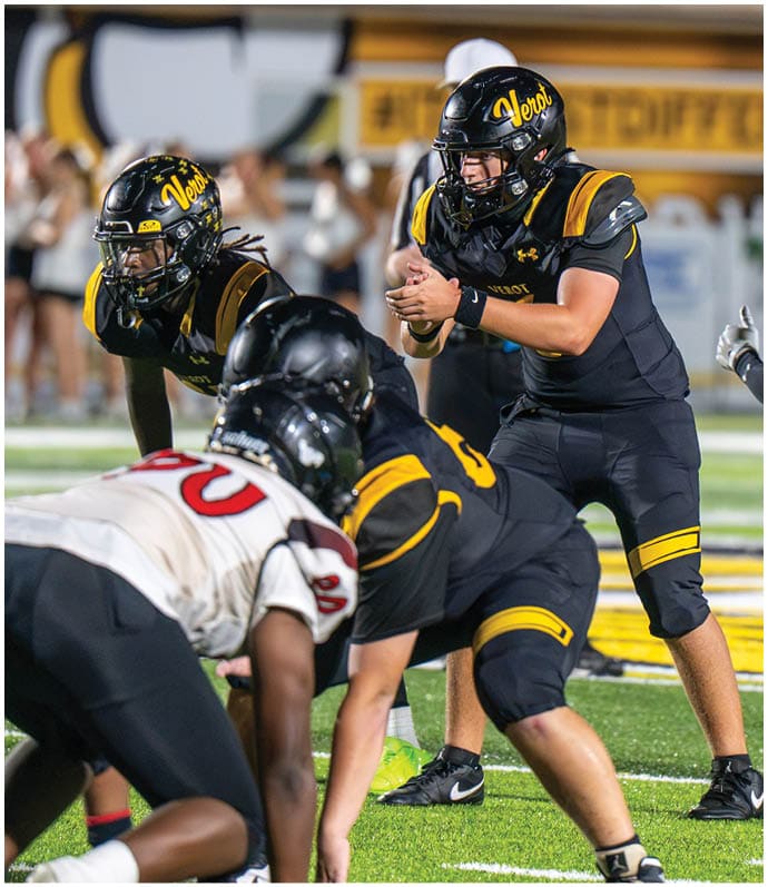 Two people in black and yellow football uniforms are positioned on a football field, with one person holding a football and the other preparing to receive it. Descripci n generada con IA