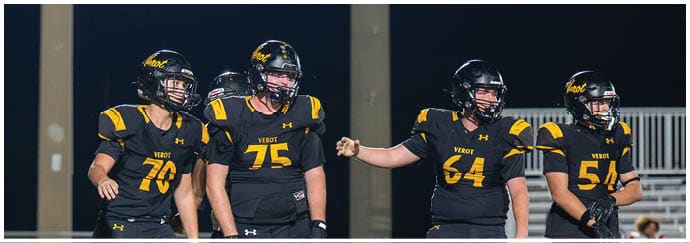 Four people wearing black and yellow football uniforms stand together on a field at night. Descripci n generada con IA