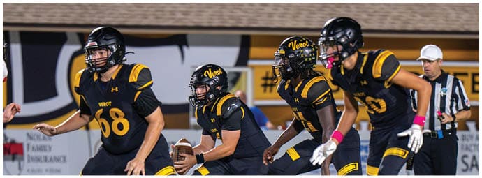 Four people in black and yellow football uniforms are on a field preparing for a play with a referee standing nearby. Descripci n generada con IA