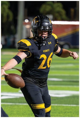 A person in a black and yellow football uniform is preparing to hand off a football to another person in a white and navy uniform on a green football field. Descripci n generada con IA