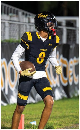 A person wearing a black and yellow football uniform with the number 8 and the word “VEROT" on the jersey is holding a football and standing on a grassy field near an orange marker. Descripci n generada con IA