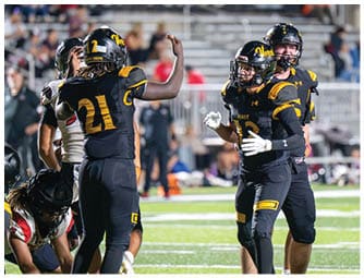 Four football players in black and yellow uniforms are on a field with one player raising an arm while others appear to be celebrating or communicating during a game. Descripci n generada con IA