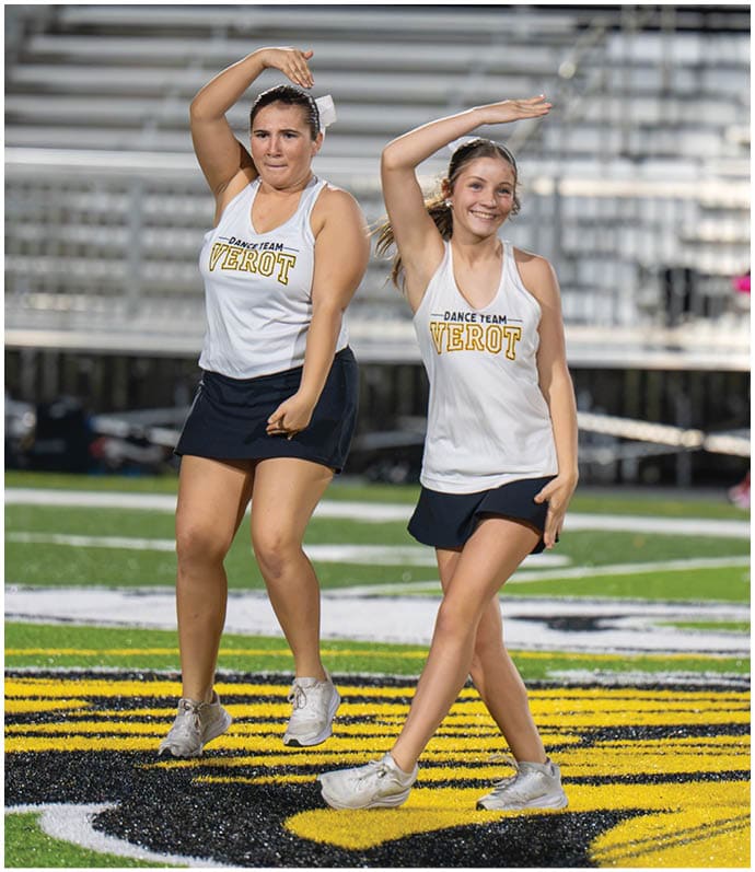 Two adults wearing matching dance team tank tops and skirts are performing a synchronized dance routine on a sports field. Descripci n generada con IA