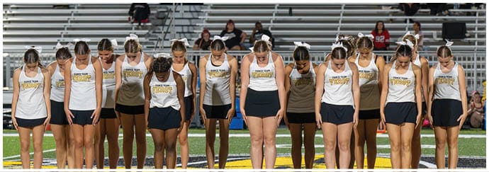 A group of people wearing matching athletic uniforms stand in a line on a sports field with their heads bowed. Descripci n generada con IA