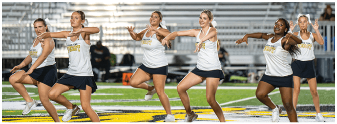 Six adults wearing matching athletic outfits perform a synchronized dance routine on a sports field with bleachers in the background. Descripci n generada con IA