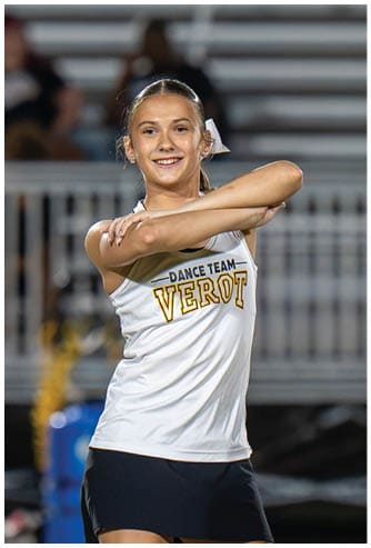A person wearing a white dance team top and black skirt is smiling with arms crossed in front of their chest in an outdoor stadium setting. Descripci n generada con IA