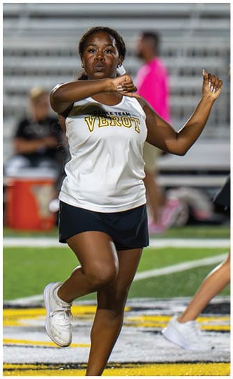 A person wearing a white sports jersey and black shorts is performing a dance or cheerleading move on a sports field with blurred spectators in the background. Descripci n generada con IA