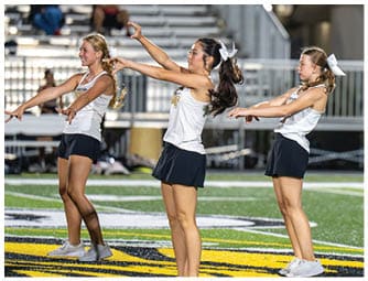 Four adults wearing white tops, black skirts, and white shoes perform a synchronized dance or cheer routine on a sports field with a large yellow and black logo. Descripci n generada con IA