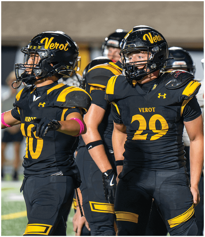 Two people in black and yellow football uniforms with helmets stand on a field, one with arms extended and the other looking to the side. Descripci n generada con IA