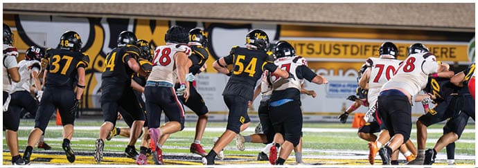 People wearing football uniforms are actively running and engaging in a play on a brightly lit football field. Descripci n generada con IA