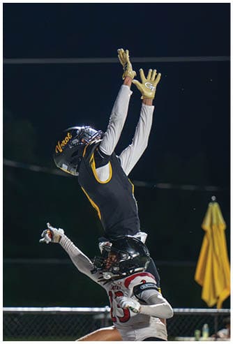 Two people in football gear are jumping and reaching for a ball during a nighttime game with a fence and seated spectators in the background. Descripci n generada con IA