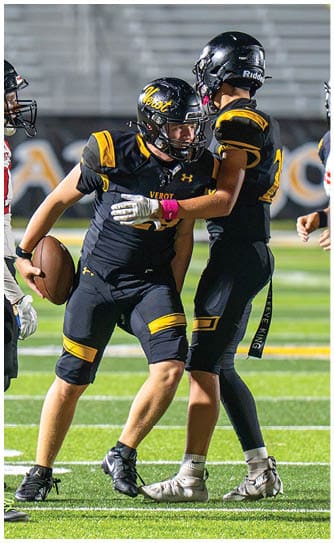 A person in a black and yellow football uniform is holding a football and appears to be preparing to run while another person in a white and red uniform stands nearby on a football field. Descripci n generada con IA