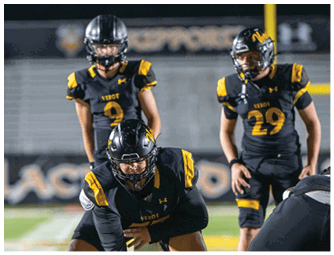 Three people wearing black and yellow football uniforms and helmets are positioned on a field, with one person crouched in the foreground and two standing behind. Descripci n generada con IA