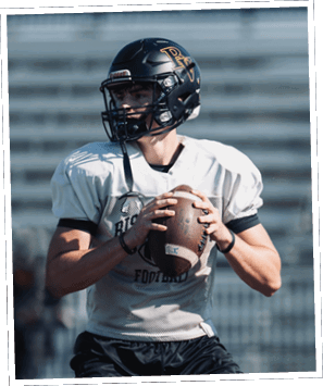 A person wearing a football helmet and uniform is holding a football in a ready-to-throw position on a field with empty bleachers in the background. Descripci n generada con IA
