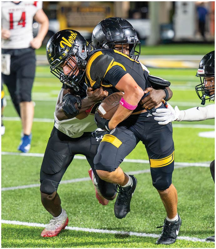 A person in black and yellow football gear is running with the ball while being tackled by two other players in black and white uniforms on a football field. Descripci n generada con IA