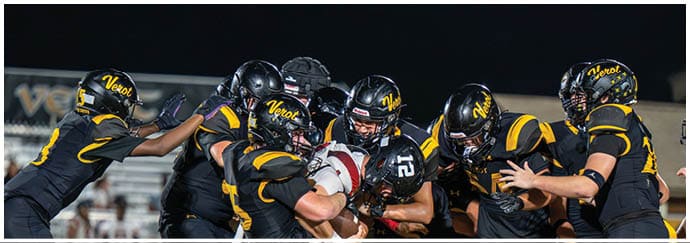 A group of football players in black and yellow uniforms are tightly clustered as they tackle and try to stop a player in a white and red uniform holding the ball during a game. Descripci n generada con IA
