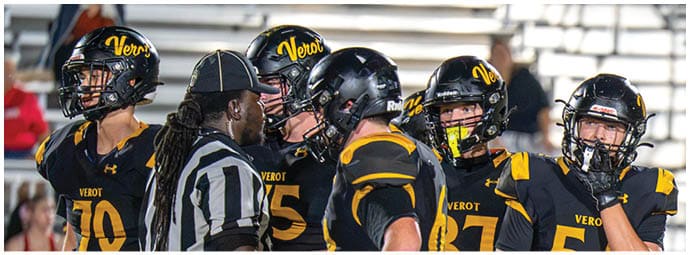 Several people wearing black and yellow football uniforms and helmets are gathered closely with a referee in a striped shirt on a sports field. Descripci n generada con IA