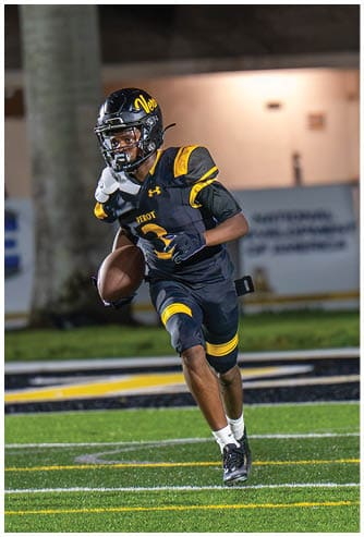 A person wearing a black and yellow football uniform is running on a field while holding a football. Descripci n generada con IA