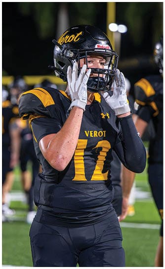 A person wearing a black and yellow football uniform with the number 12 and a helmet is adjusting their gloves on a football field with other players in the background. Descripci n generada con IA