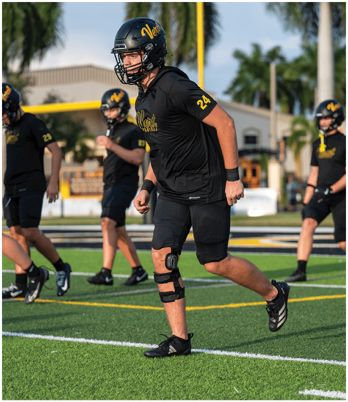 A person wearing a black football uniform and helmet with the number 24 is running on a football field with other players in the background. Descripci n generada con IA