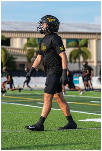 A person wearing a black sports uniform with the number 22 and a helmet is walking on a green football field with other players and a building in the background. Descripci n generada con IA