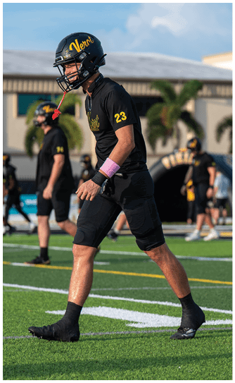 An adult wearing a black football uniform with the number 23 and a helmet is walking on a green football field with other players and a building in the background. Descripci n generada con IA